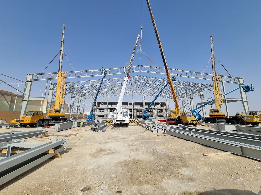 Mobile cranes and boom lifts raising a roof truss across the hall's 64 m clear span