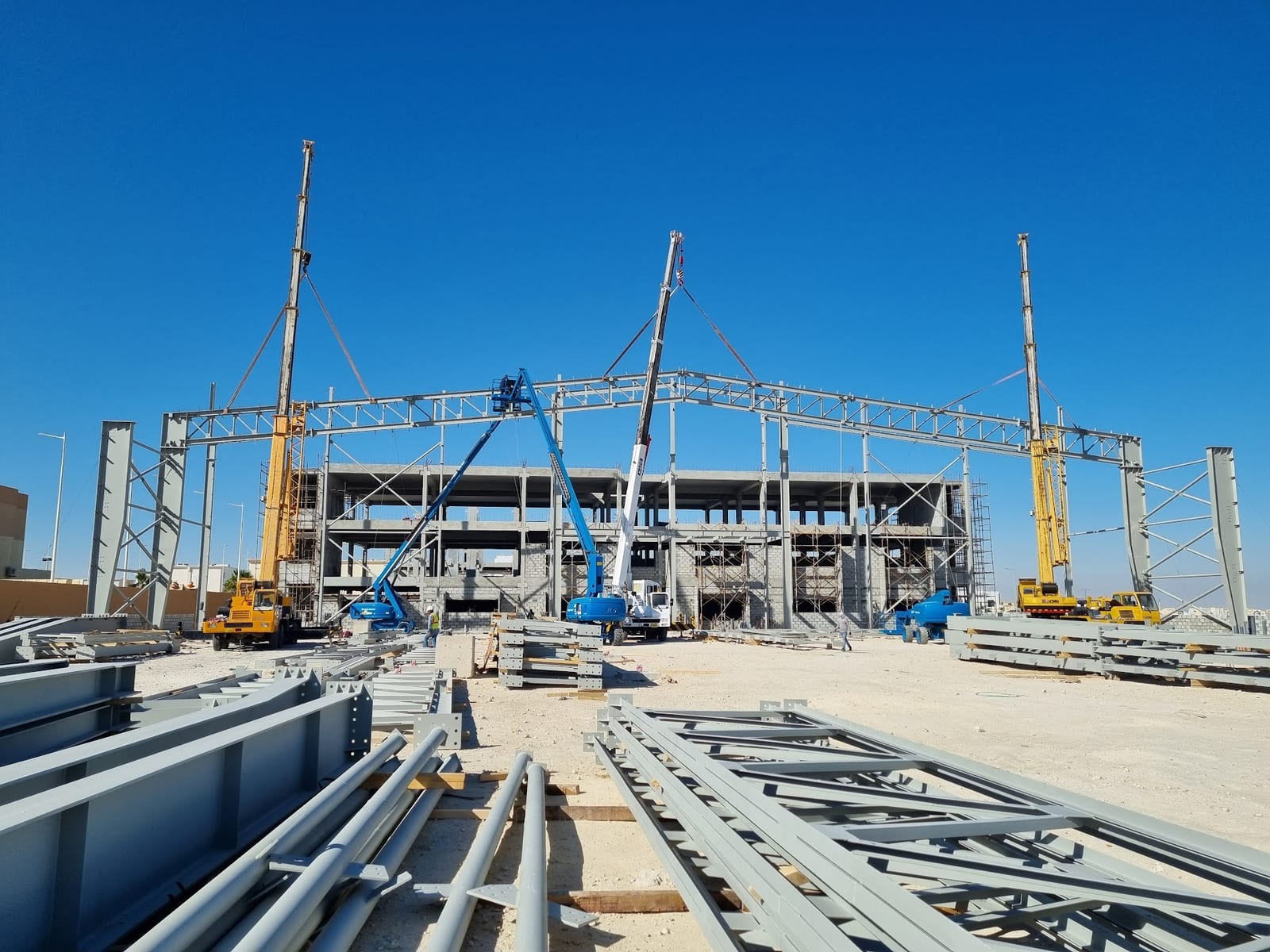 Rafters and lattice sections laid out on the sand beside the part-erected frame and its standing cranes